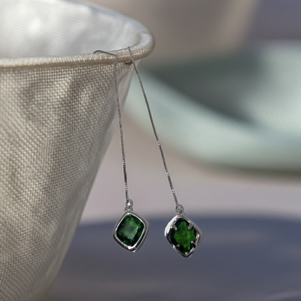 A pair of silver dangle earrings with square-cut green gemstones, hanging from a textured beige fabric holder against a soft, blurred background.