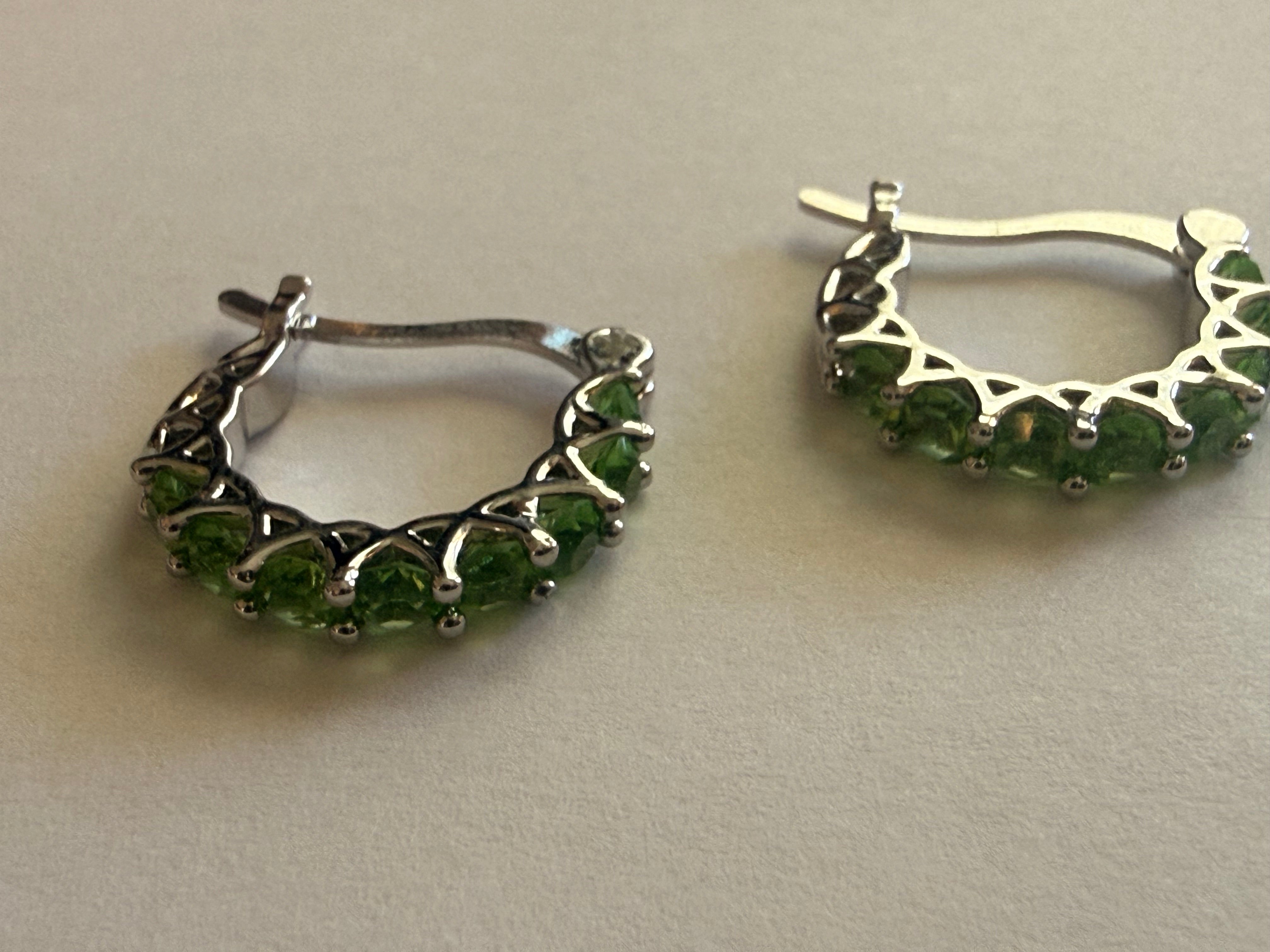 Silver-tone hoop earrings set with a row of round green rhinestones in prong settings, openwork triangular gallery, latch-back closure, angled view