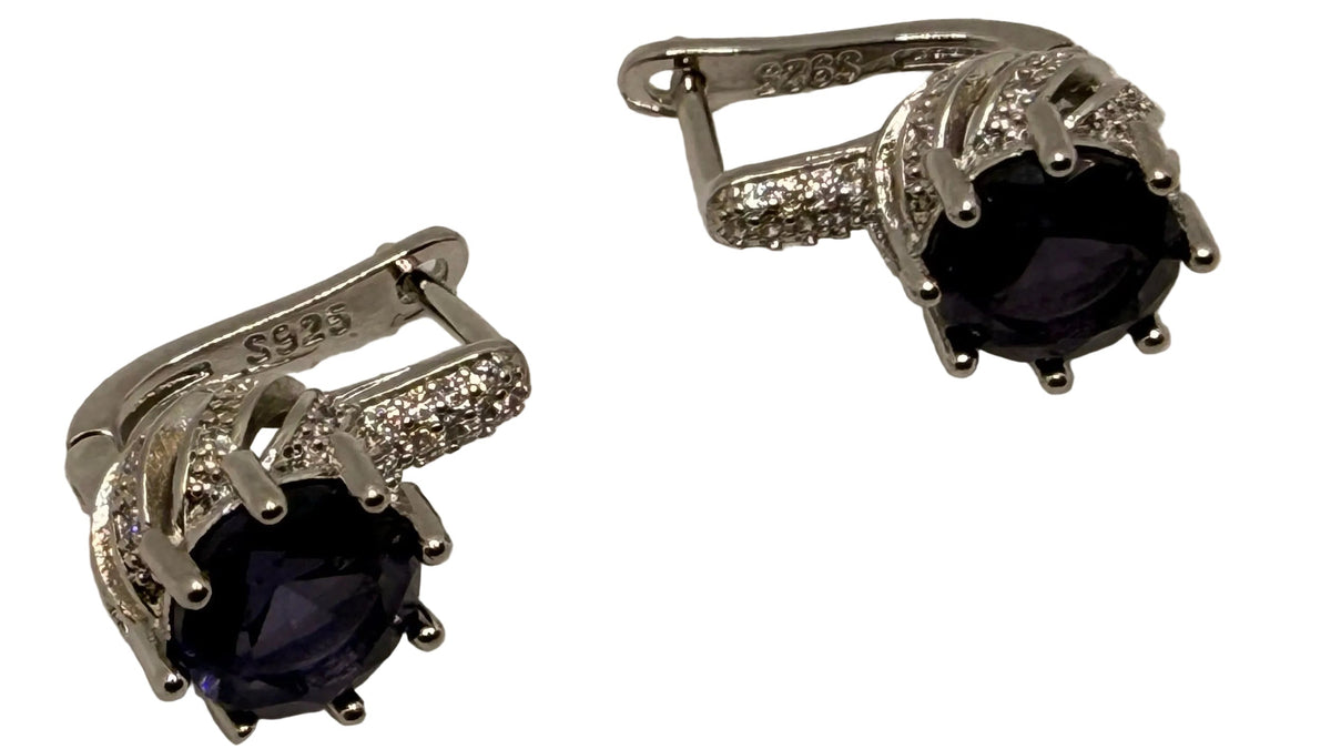 A pair of silver-toned huggie hoop earrings with oval dark purple gemstones and small clear accent stones, displayed on a textured white surface.